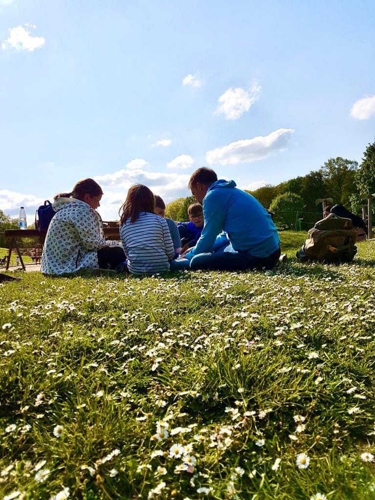 Kinder und ein Betreuer sitzen bei Sonnenschein auf einer Wiese voller Gänseblümchen