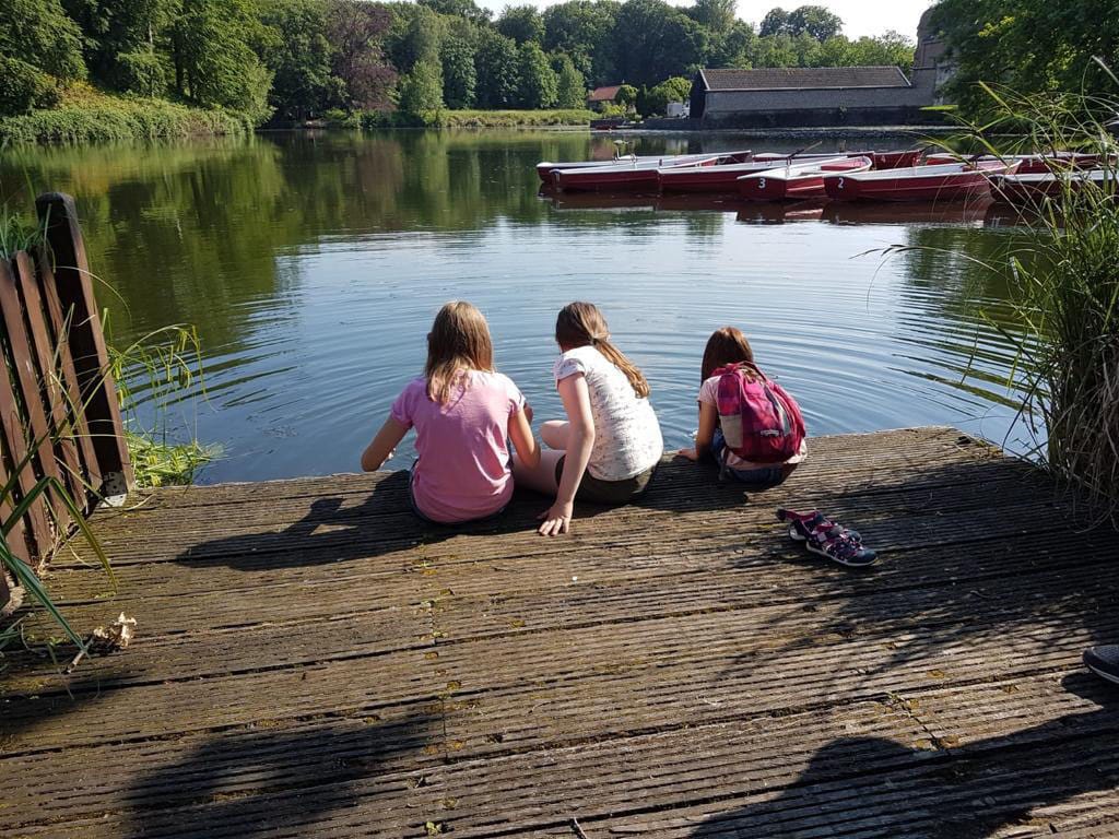 Drei Mädchen sitzen nebeneinander auf einem Holzsteg und schauen auf einen See mit Tretbooten