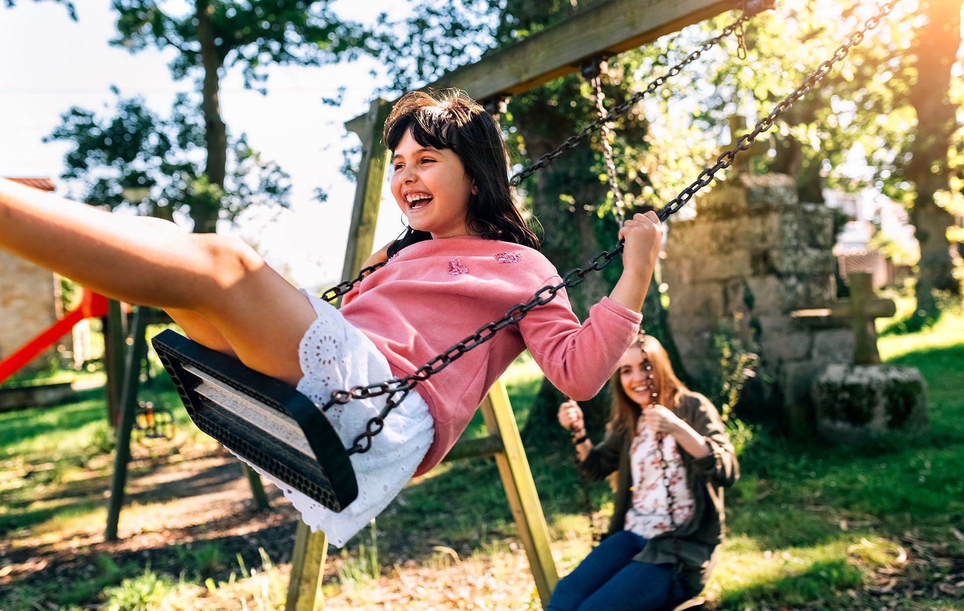 Mädchen lacht ausgelassen auf einer Schaukel, im Hintergrund eine Betreuerin